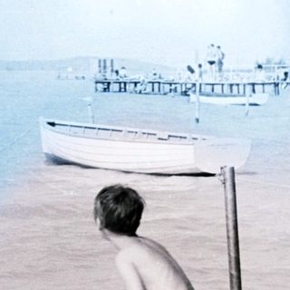 Boy sits on dock in water, boats and shoreline visible.