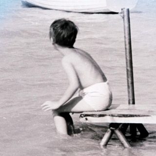 Boy sits on dock in water, boats and shoreline visible.
