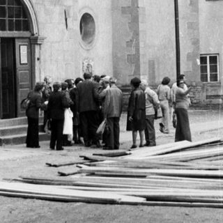 People gather outside a stone building with a tower.