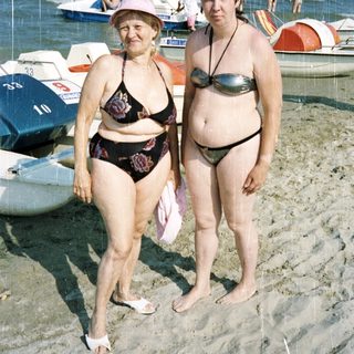 Two women stand on a beach with boats in the background.