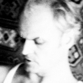 Man reading newspaper, woman seated on patterned couch.