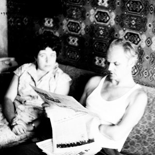 Man reading newspaper, woman seated on patterned couch.