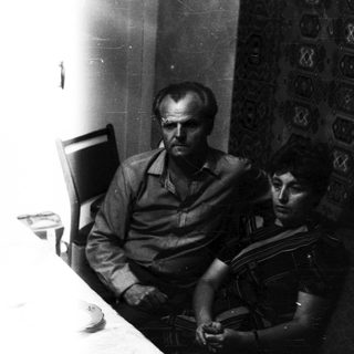 Here's a brief summary of the image: Two men seated, looking down, against a patterned wall.