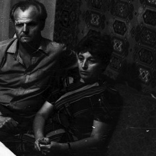 Here's a brief summary of the image: Two men seated, looking down, against a patterned wall.