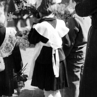 Children in uniforms present flowers to women outside a building.