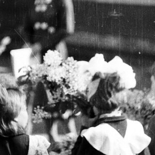Children in uniforms present flowers to women outside a building.