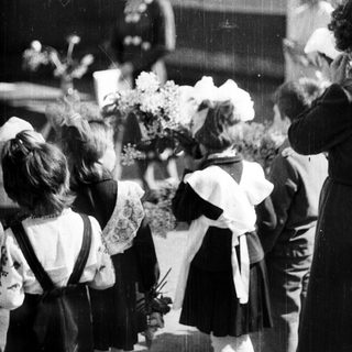 Children in uniforms present flowers to women outside a building.