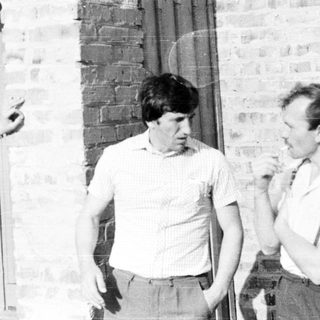 Here's a brief summary of the image: Three men stand outside a brick building, one smoking.