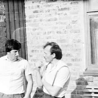 Here's a brief summary of the image: Three men stand outside a brick building, one smoking.