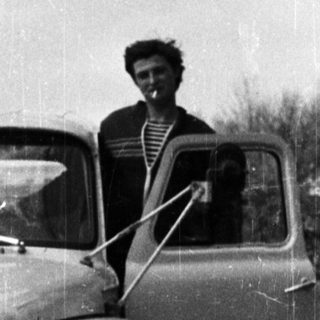 Man sitting in vintage truck, door open, black and white.