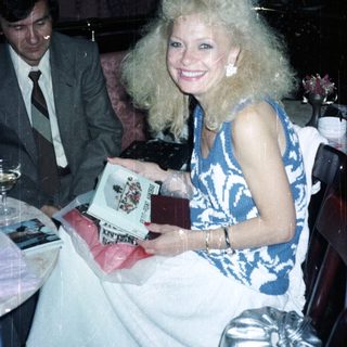 Woman with large hair holds gift, man nearby.