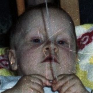 Baby sits in crib with toy and patterned blanket.