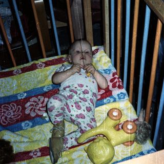 Baby sits in crib with toy and patterned blanket.
