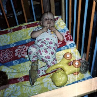 Baby sits in crib with toy and patterned blanket.