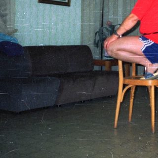 Here's a brief summary of the image: Man seated, others balance bottles on chairs in a room.