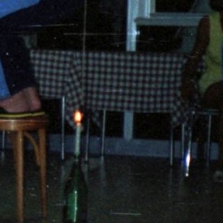 Here's a brief summary of the image: Man seated, others balance bottles on chairs in a room.