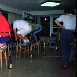 Here's a brief summary of the image: Man seated, others balance bottles on chairs in a room.