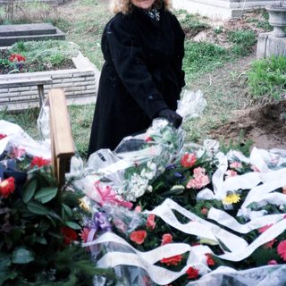 Woman stands by a grave, surrounded by flowers and ribbons.