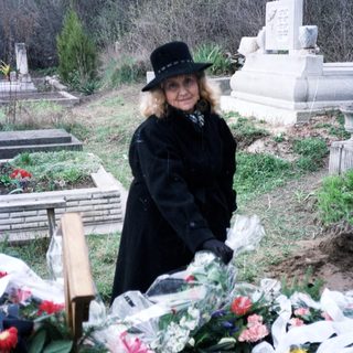 Woman stands by a grave, surrounded by flowers and ribbons.