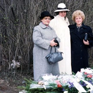 Here's a brief summary of the image: Women stand near a grave adorned with flowers.