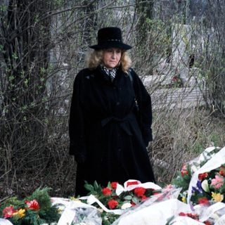Woman stands near a grave adorned with flowers and plaque.