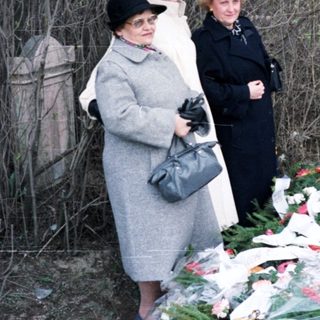 Here's a brief summary of the image: Women stand near a grave covered with flowers.