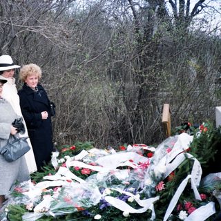 Here's a brief summary of the image: Women stand near a grave covered with flowers.