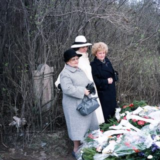 Here's a brief summary of the image: Women stand near a grave covered with flowers.