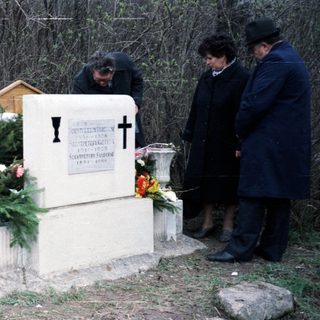 People visit a gravesite with a cross and flowers.