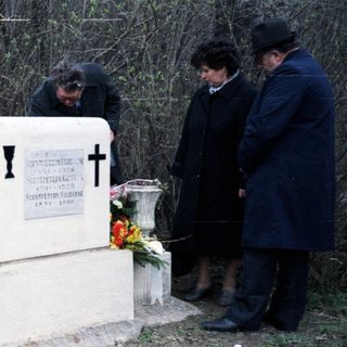 People visit a gravesite with a cross and flowers.