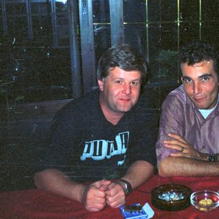 Two men sit at a table with drinks and ashtrays.