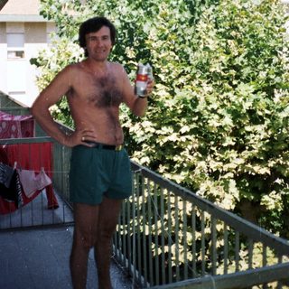 Man stands shirtless on a balcony, holding a beer.