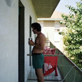 Man stands on balcony holding a drink, laundry drying.