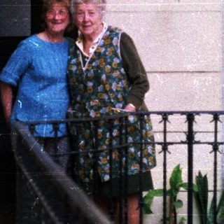 Two women stand on a balcony with potted plants.