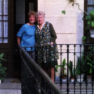 Two women stand on a balcony with potted plants.