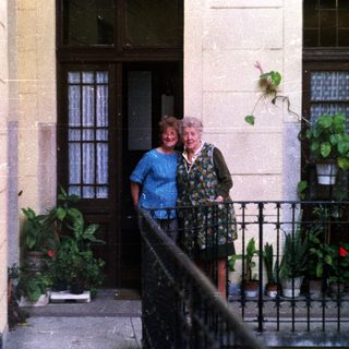 Two women stand on a balcony with potted plants.