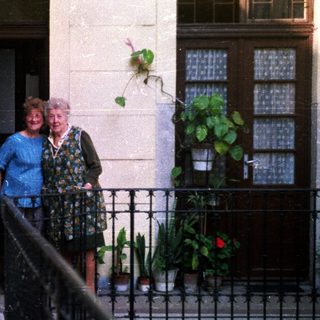 Two women stand on a balcony with potted plants.