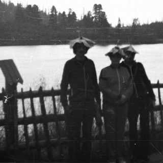 Three people stand near a body of water, wearing rain hats.