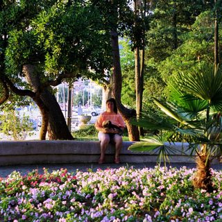 Woman sits near flowers and boats in a coastal scene.