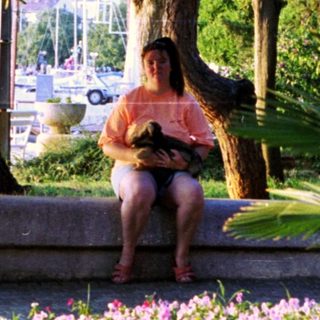 Woman sits near flowers and boats in a coastal scene.