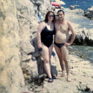 Couple standing on a rocky beach with people sunbathing.