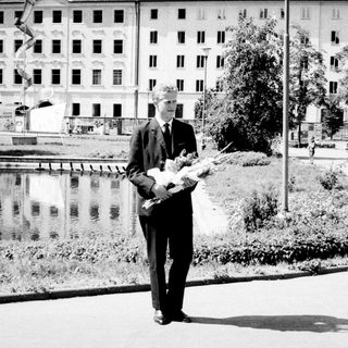 Man in suit holds flowers, standing near a body of water.