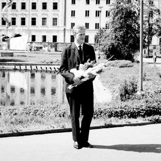 Man in suit holds flowers, standing near a body of water.