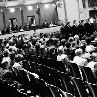 Group of men on stage, audience seated in tiered theater.