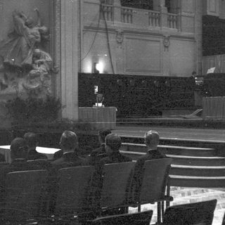 Men seated, observing a stage with a statue.