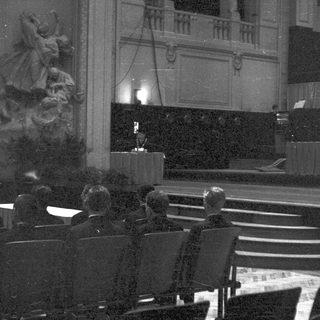 Audience seated before a stage with a statue visible.