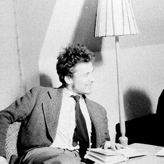 Two people converse near books and a lamp.