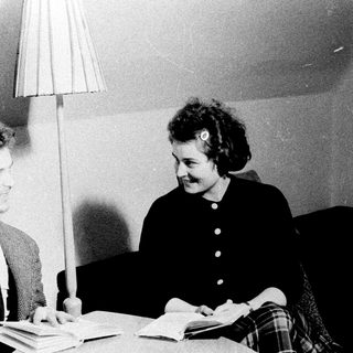 Two people converse near books and a lamp.