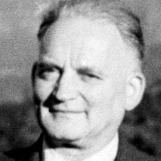 Man in suit stands near pine trees with a hill backdrop.
