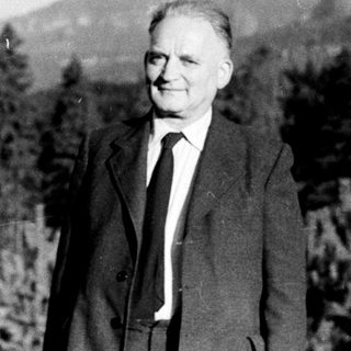 Man in suit stands near pine trees with a hill backdrop.
